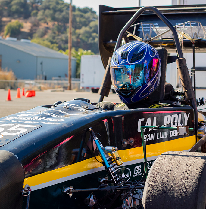 A driver wearing a blue helmet sitting in the Cal Poly Racing Formula car on a sunny day.