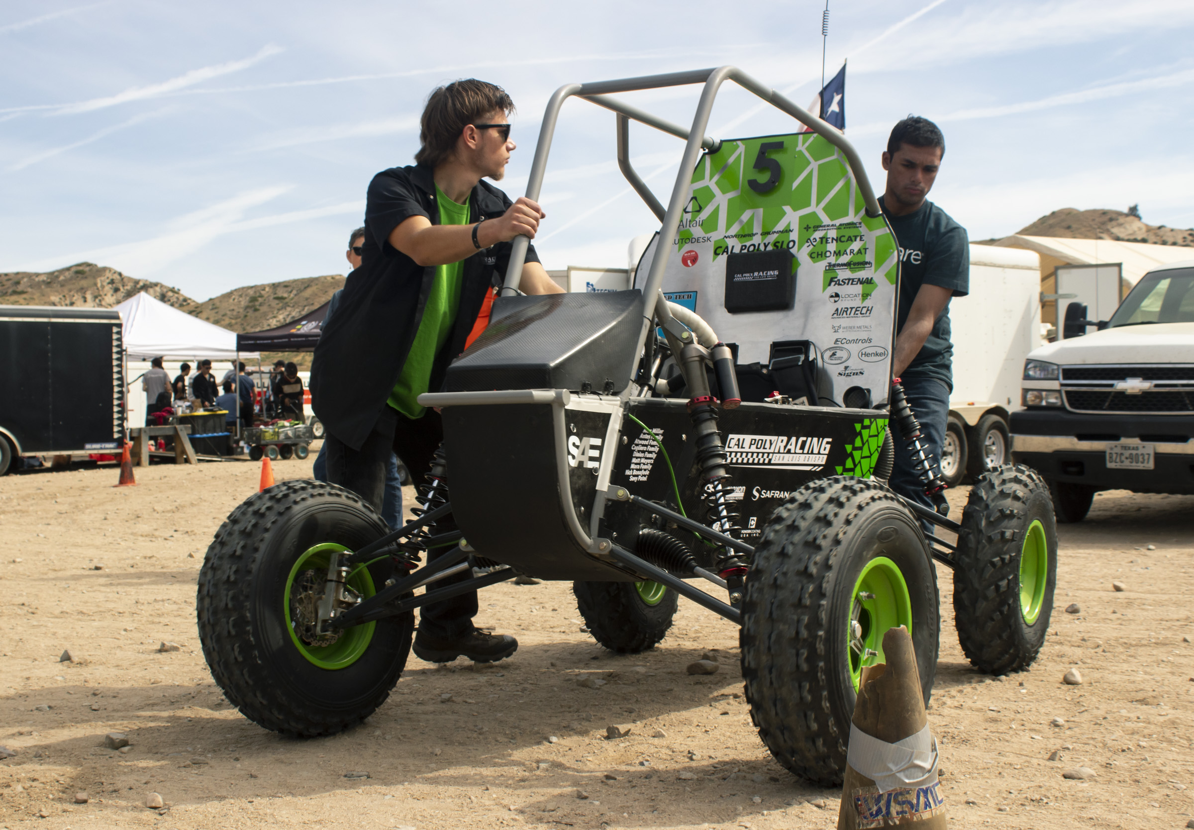 Two men roll the Cal Poly Racing Baja car on a dirt plain. There are pop-up tents, trailers, and pick-up trucks in the background.