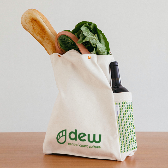 An off-white cloth shopping bag, decorated with the Dew logo and vine graphic, containing a loaf of bread, lettuce, and a wine bottle.