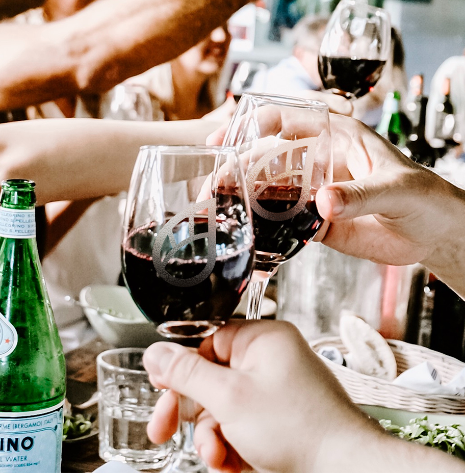 A close up shot of two wine glasses, each engraved with the Dew logo, during a toast at a lively, crowded table.