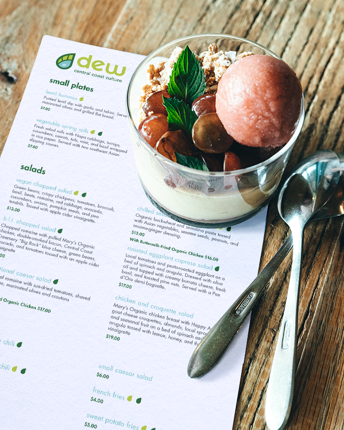 A Dew menu on a wooden table underneath a parfait dessert and two spoons.