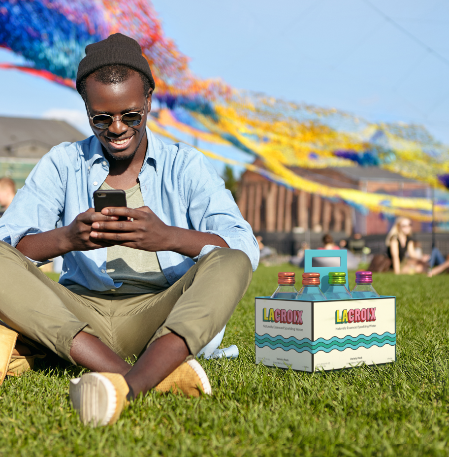 A man sits cross-legged in a park texting his friends. A six-pack case of LaCroix is on the grass next to him and colorful streamers fly behind him.