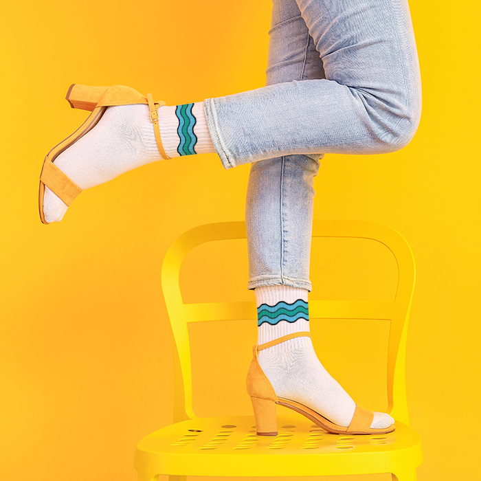 A woman, shown from the thighs down, stands on a yellow chair in a yellow room. She wears white socks with a stylized water graphic around the ankles.