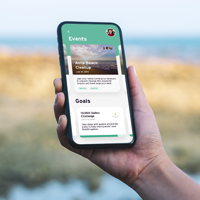 A hand holding a phone at the beach. The screen shows Loop community events for a beach cleanup and a water-saving challenge.